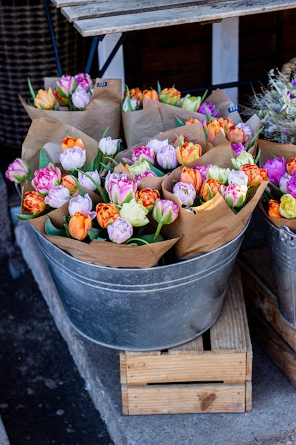 A large galvanized metal tub filled with dozens of freshly cut tulips in vibrant shades of pink, orange, yellow, and purple, each wrapped in brown craft paper. The tulips have bright green stems and l