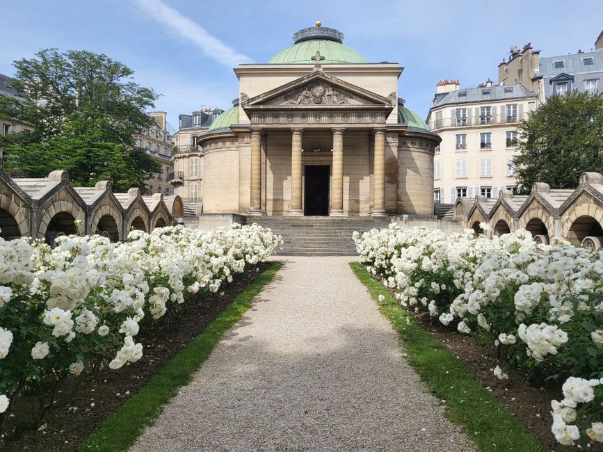 A symmetrical pathway leads to a classical stone building with a green domed roof and grand columns at the entrance, surrounded by rows of lush white roses in full bloom on both sides. The roses have 