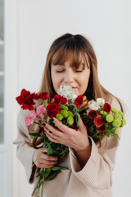 A young woman with light brown hair and bangs holds a vibrant bouquet of mixed fresh flowers close to her face, with her eyes gently closed and a subtle smile. The bouquet includes red carnations, pin