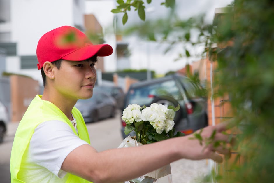 A young floristry delivery person, wearing a bright red cap and a neon yellow safety vest over a white t-shirt, is seen placing a bouquet of fresh, white hydrangeas wrapped in clear plastic against a 